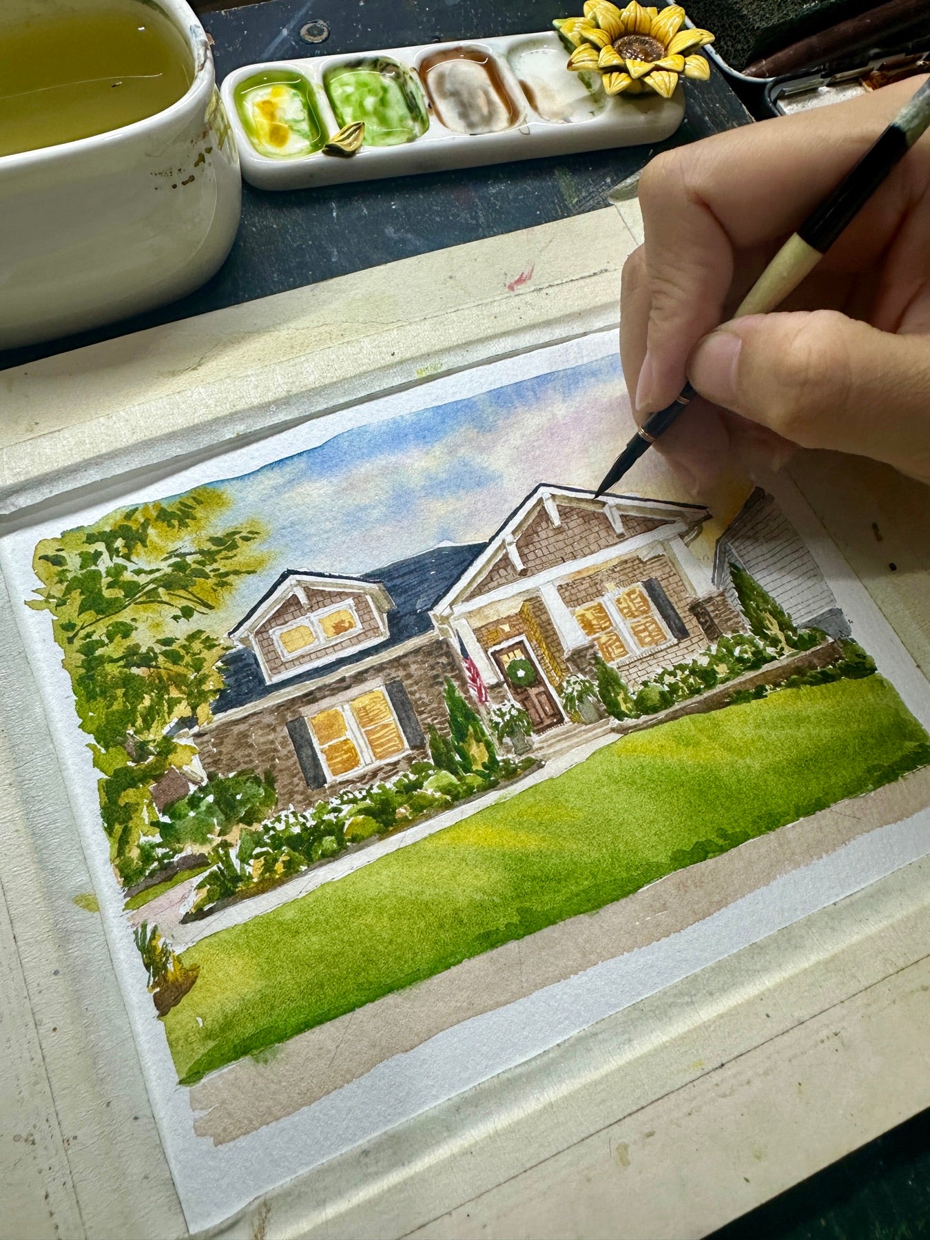 Artist hand-painting a watercolor portrait of a warm-lit family home
