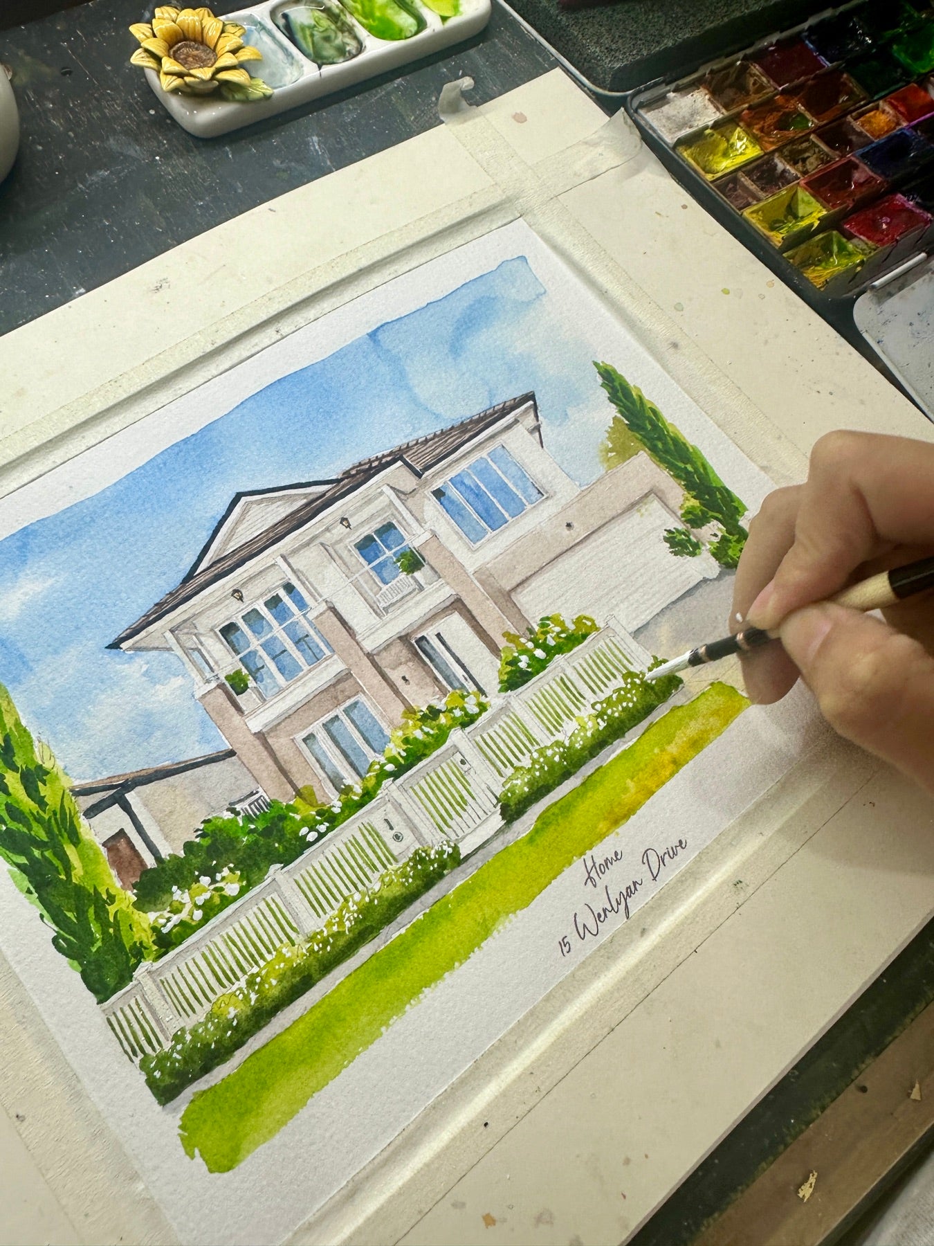 Artist hand-painting a watercolor portrait of a two-story home behind a white fence