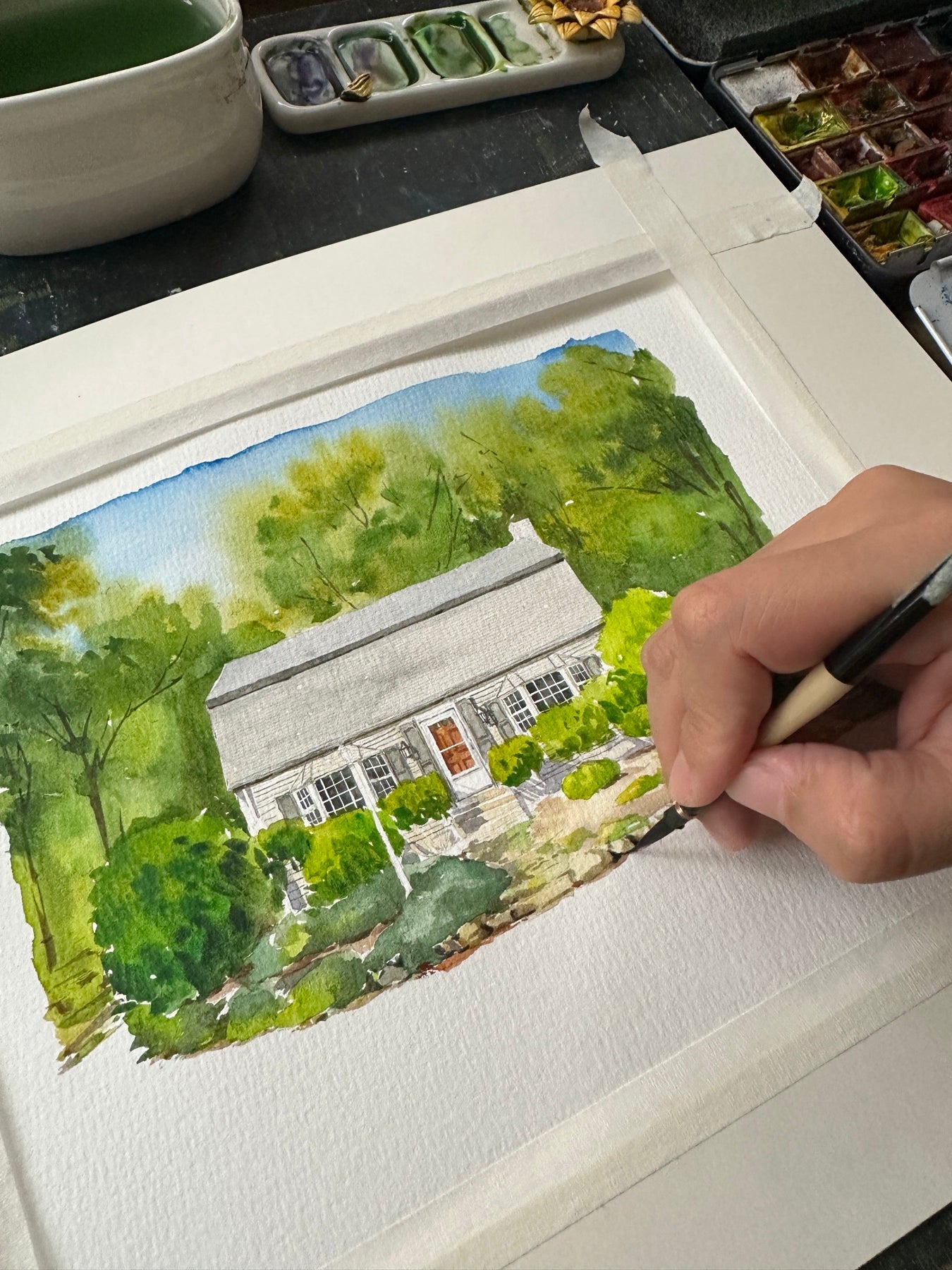 Artist hand-painting a watercolor portrait of a woodland cottage